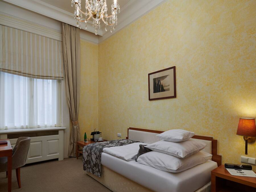 Une chambre d’hôtel avec un lit, une table de chevet, une lampe, une fenêtre avec des rideaux, un radiateur et un tableau au mur.