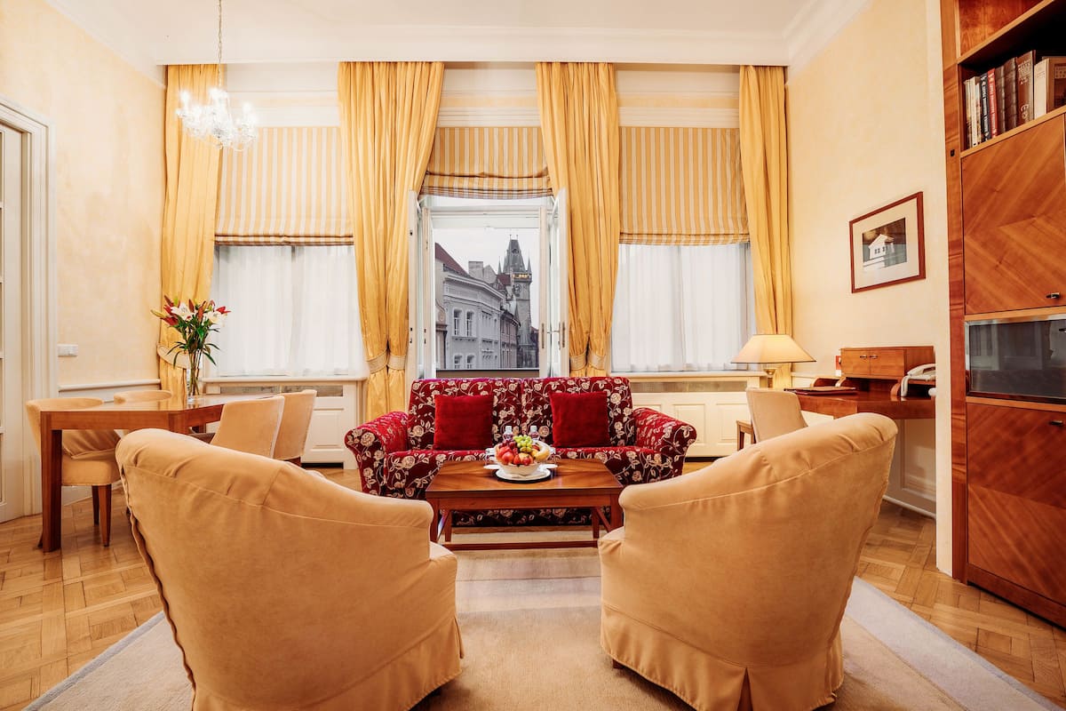Sala de estar con sofá, dos sillones, una mesa de centro con fruta, una mesa de comedor y vista a un edificio a través de cortinas.