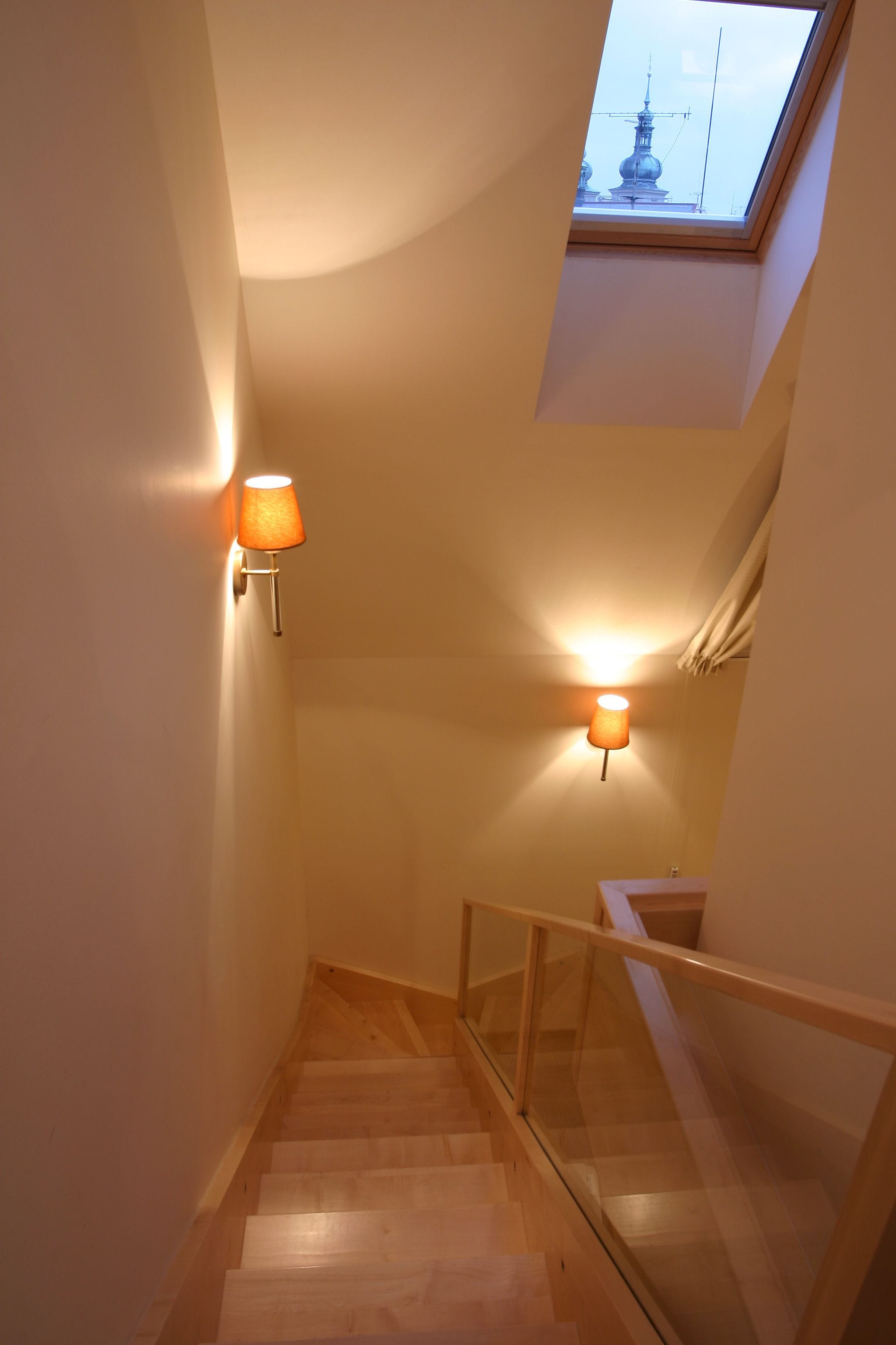 Un escalier étroit avec des marches en bois et des rampes en verre, menant à un palier surmonté d’une lucarne.