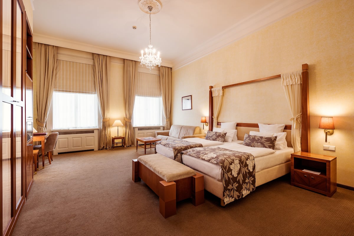 Habitación de hotel con dos camas, zona de estar, mesa de comedor y un amplio ventanal con cortinas.
