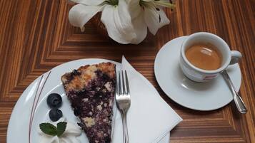 A slice of blueberry pie on a white plate with blueberries and whipped cream, a cup of coffee, and a white flower arrangement on a wooden table.