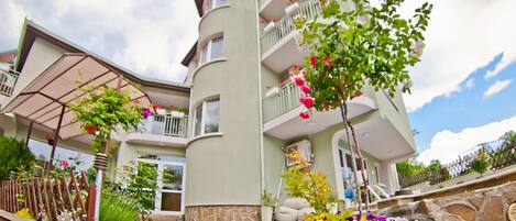 A multi-story building with a stone facade, balconies, and a garden area with flowers.