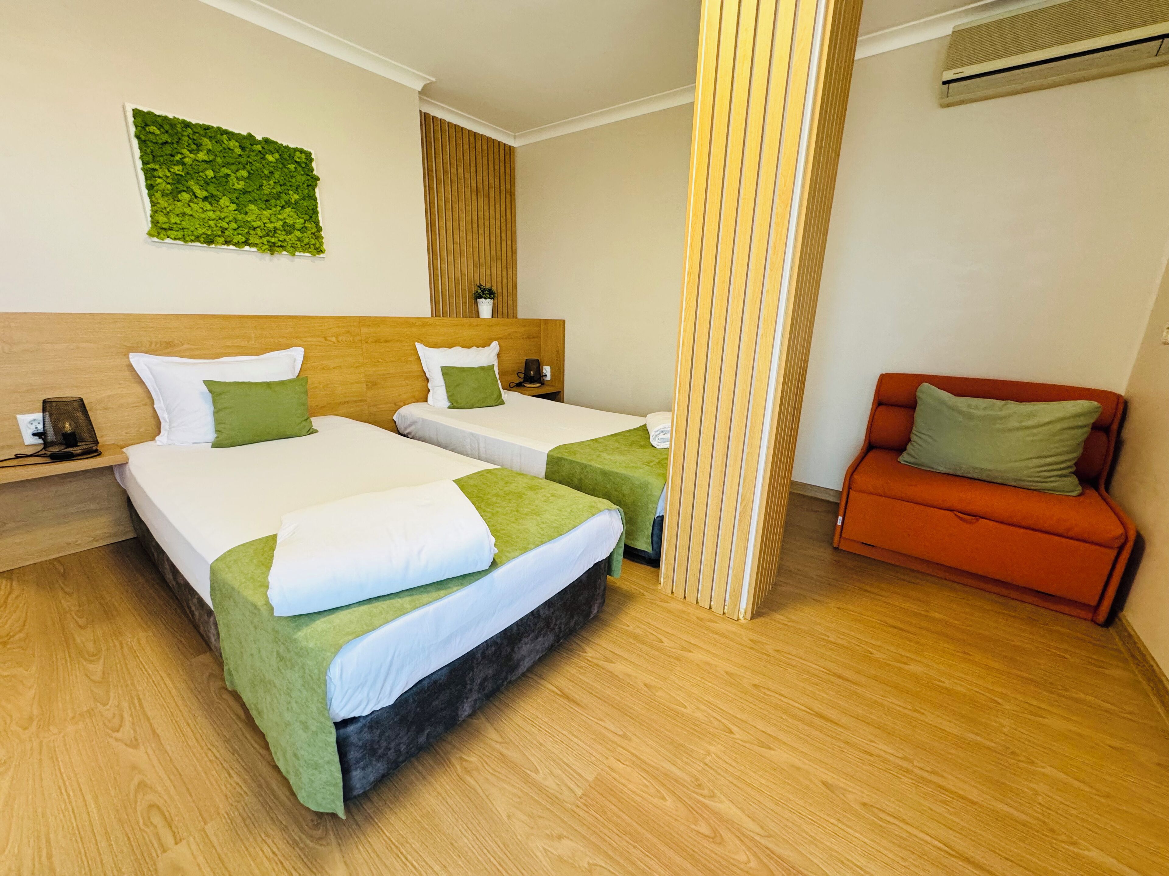 Habitación de hotel con dos camas, cabecera de madera, decoración verde en la pared, separador de madera y sofá rojo.