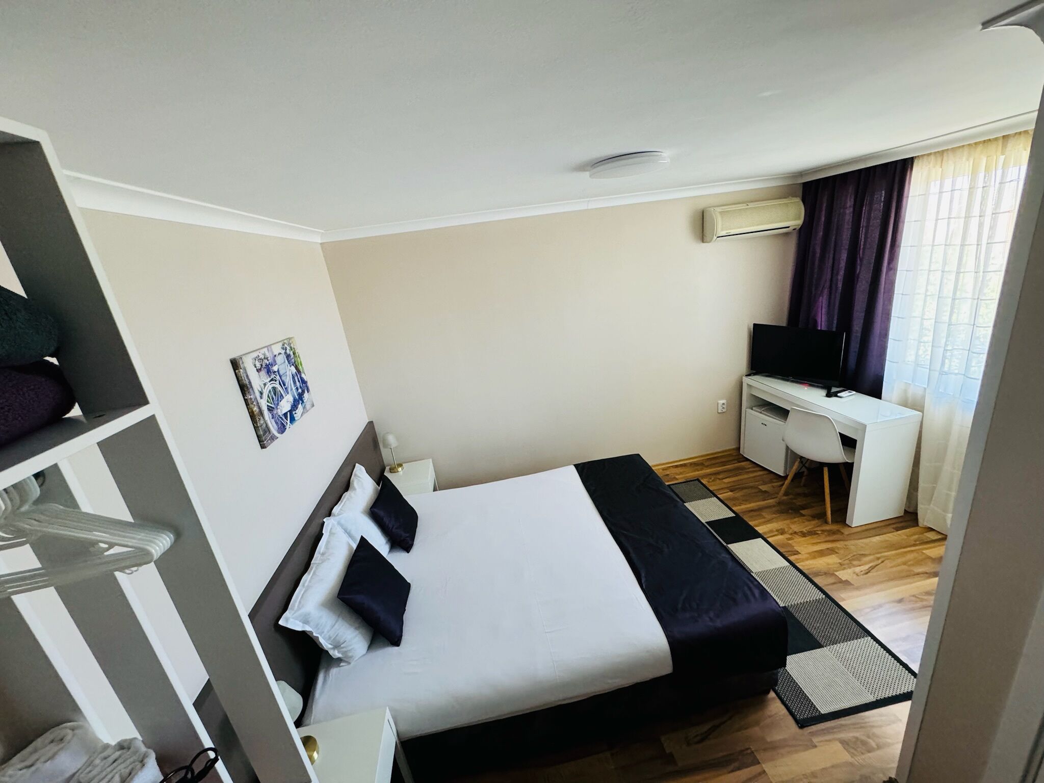 Una habitación de hotel con cama, escritorio, televisor y ventana con cortinas.