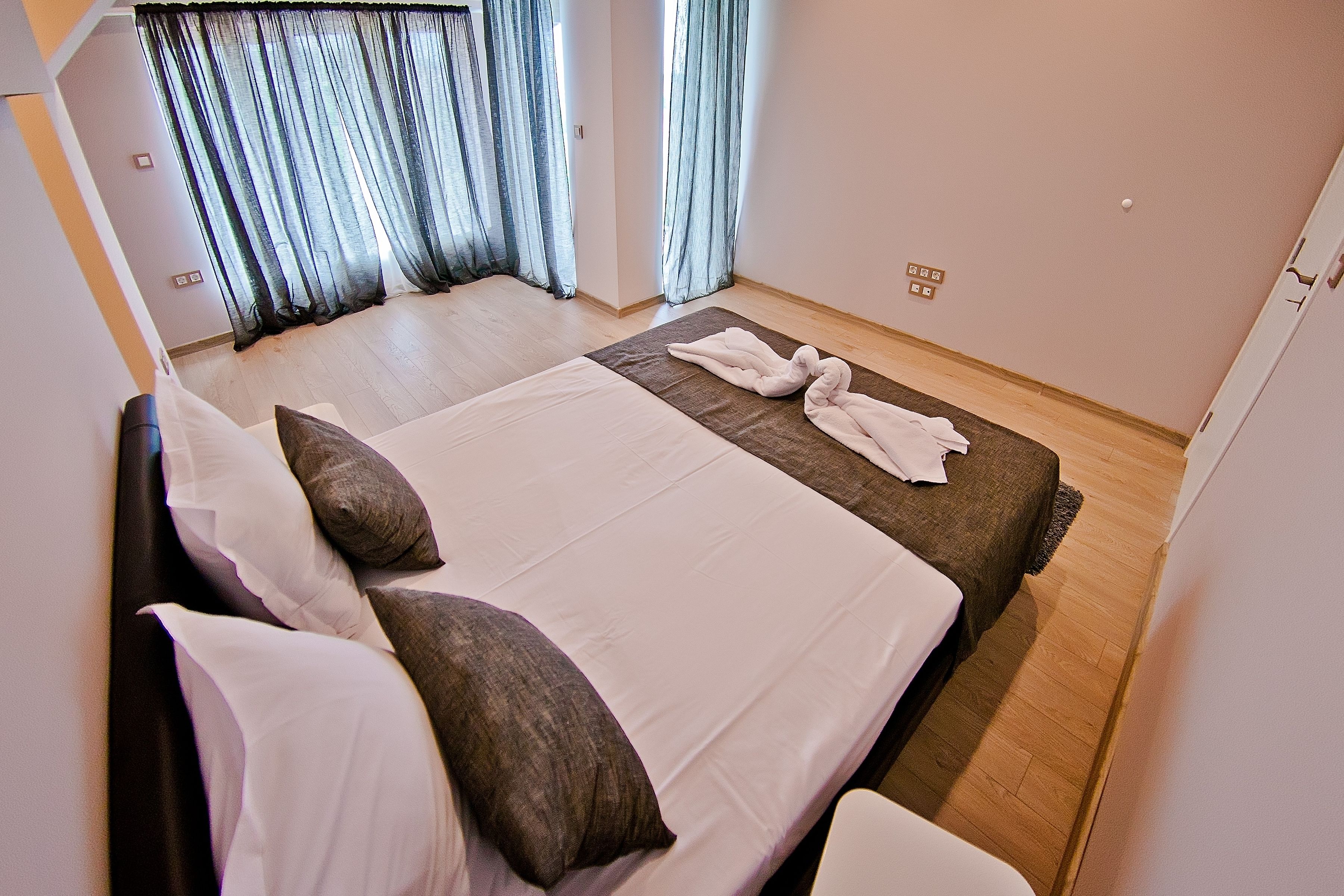 A neatly made bed with white and brown bedding, a wooden floor, and a window with sheer curtains.