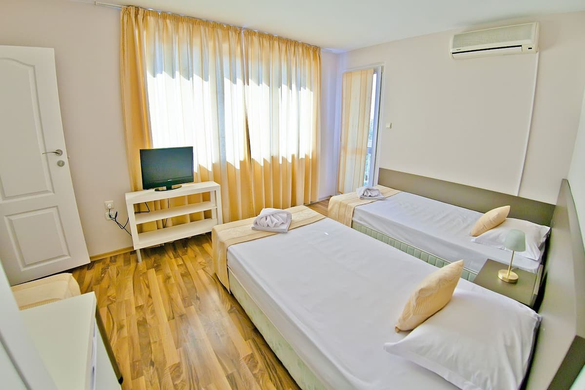 A hotel room with two beds, a TV on a stand, a window with curtains, and an air conditioning unit.