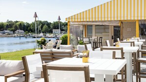 An outdoor seating area with white tables and chairs, overlooking a lake with boats.