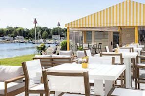 An outdoor seating area with white tables and chairs, overlooking a lake with boats.