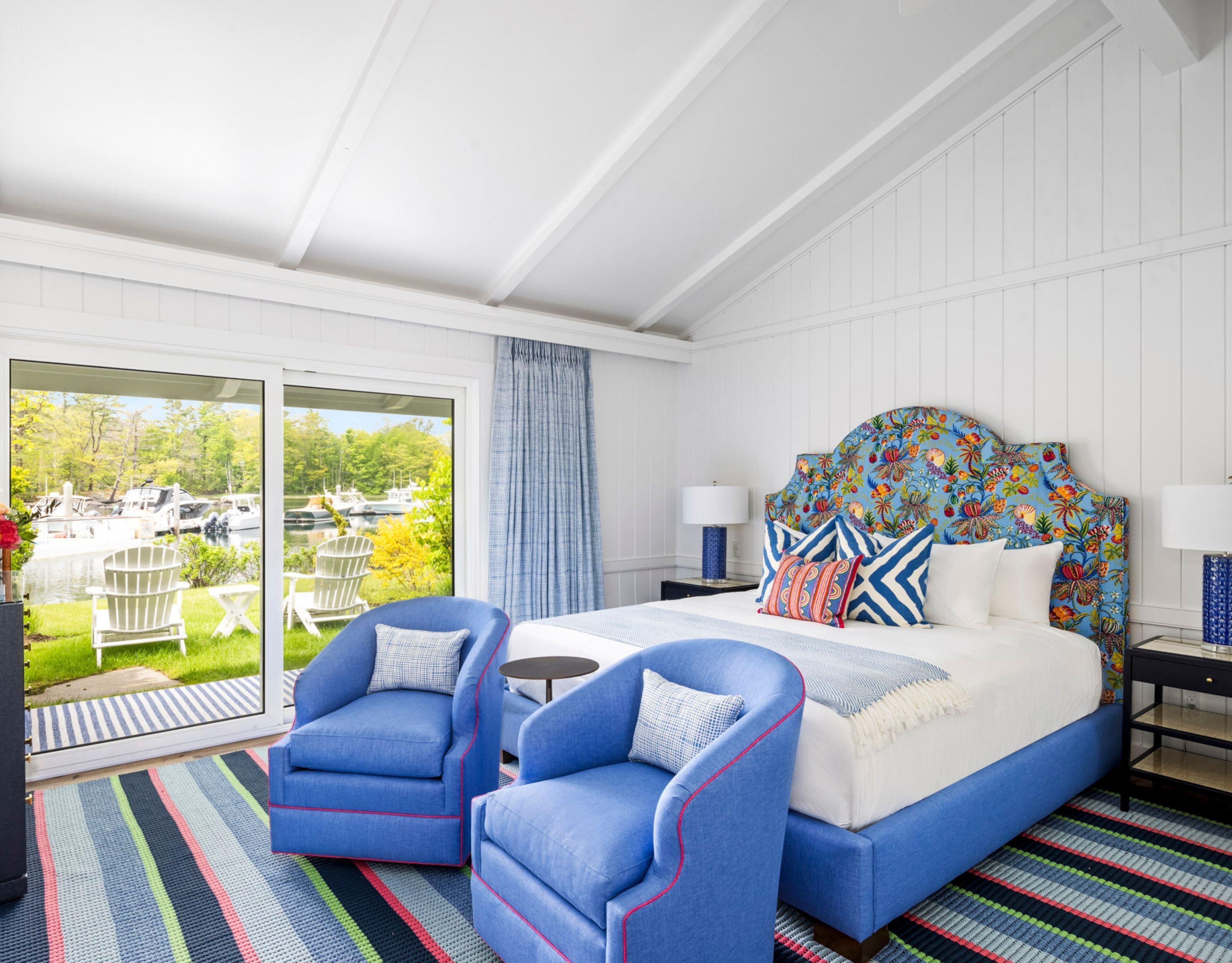 Een slaapkamer met een bed, twee blauwe fauteuils, een nachtkastje, een lamp en uitzicht op boten door een schuifglazen deur.