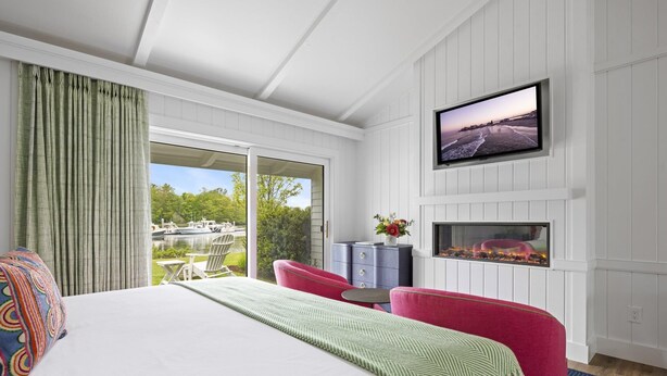 A hotel room with a bed, a TV mounted on the wall, a fireplace, and a view of boats and greenery outside.