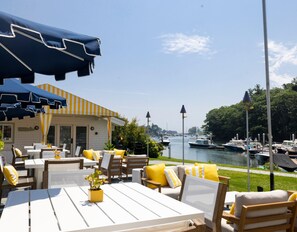 An outdoor seating area with tables and chairs, a view of the water, and a building with a yellow and white striped awning. - Yachtsman Hotel & Marina Club (Kennebunkport)