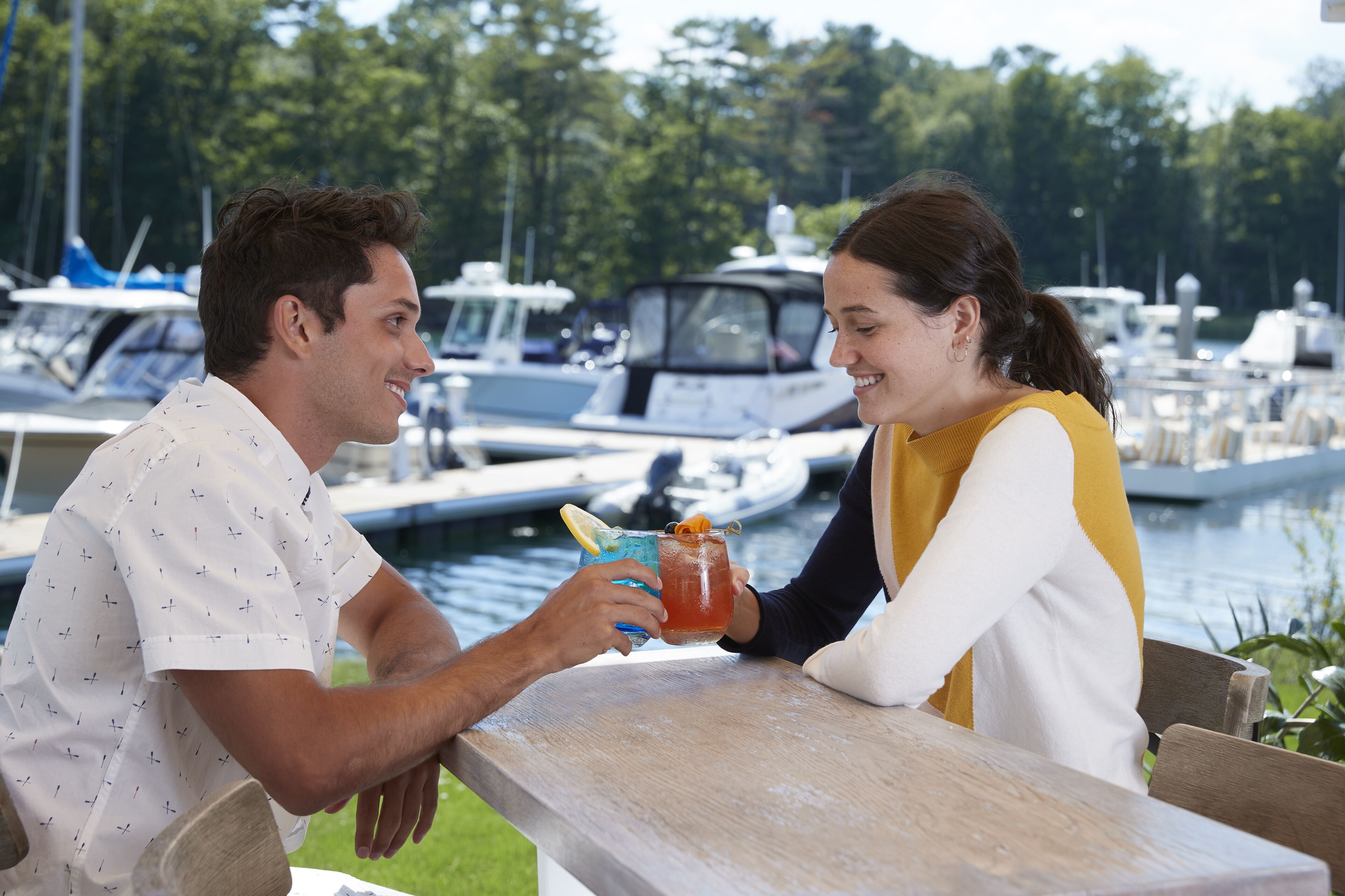 Dos personas sentadas en una mesa de madera junto a un puerto deportivo con barcos al fondo.