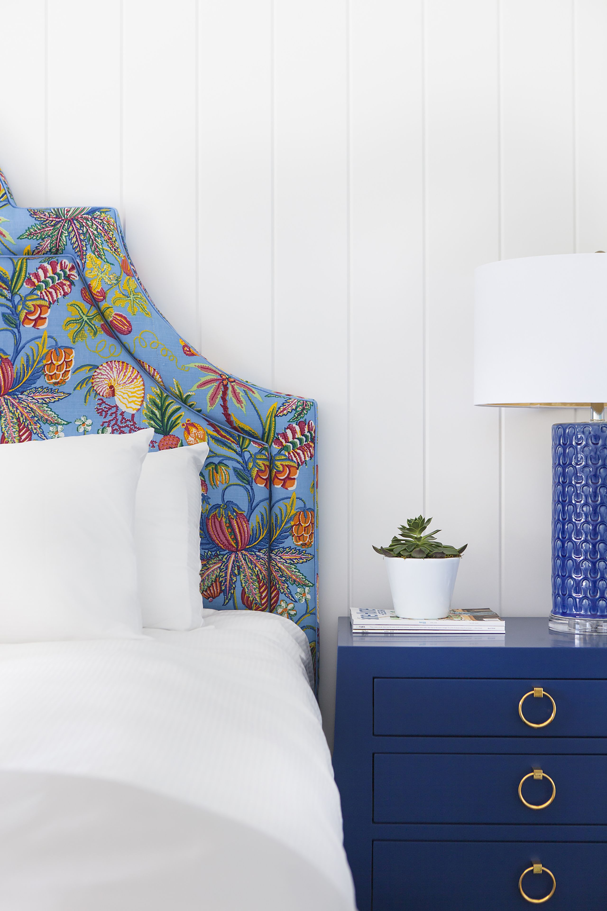 A bedroom with a floral headboard, a white bedspread, a blue nightstand with a lamp, and a small potted plant.