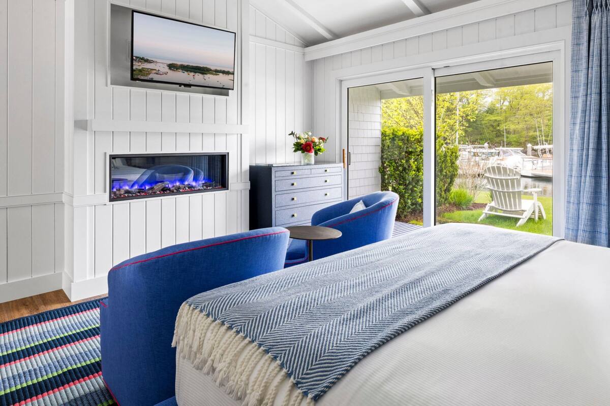 A bedroom with a bed, two blue chairs, a fireplace, a TV, and a view of a garden and outdoor seating.