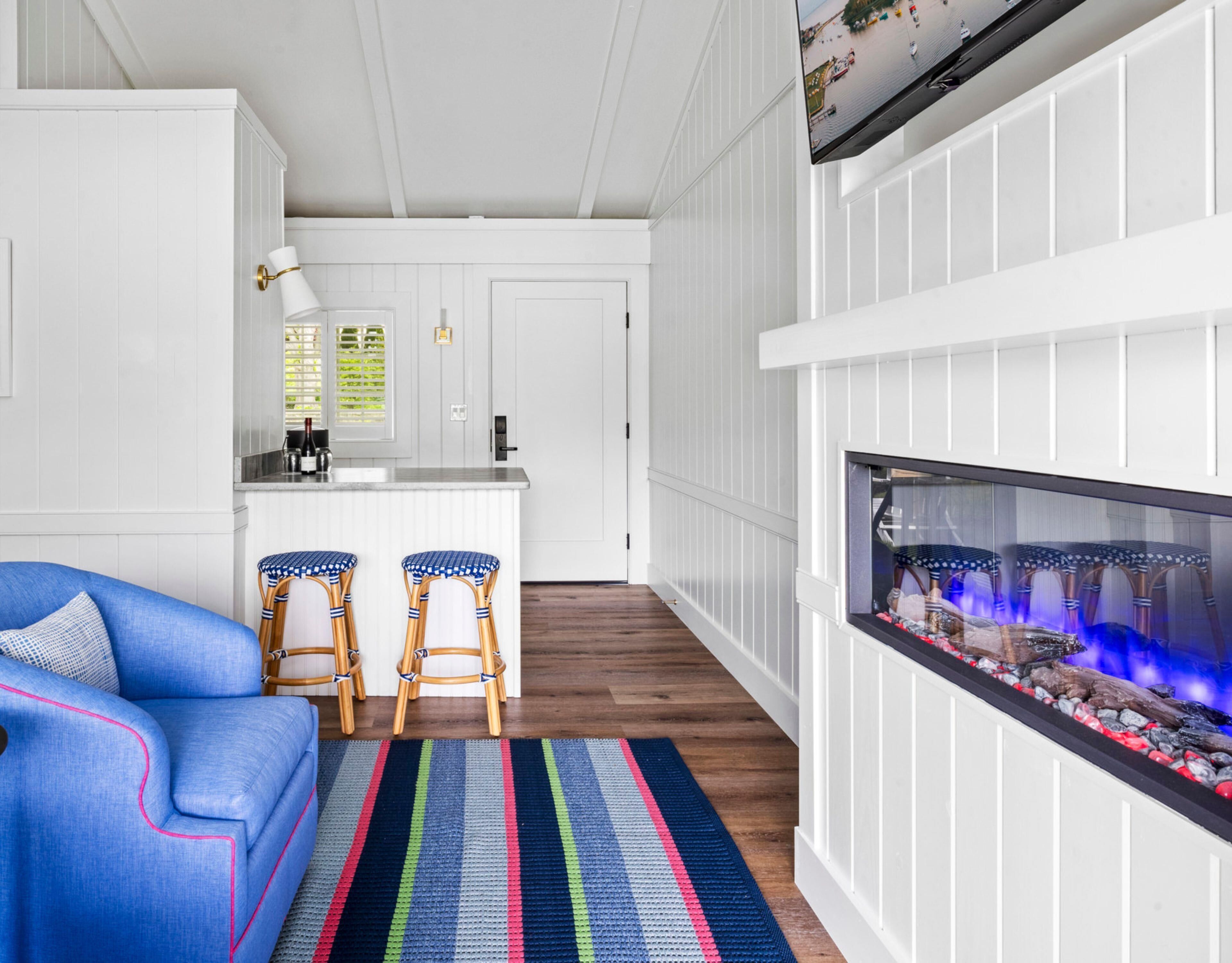 A modern living room with a blue sofa, two wicker chairs, a colorful striped rug, a fireplace, and a kitchen area with white cabinetry and a window with blinds.