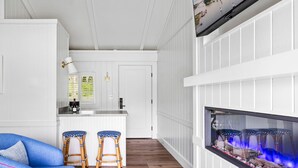 A modern living room with a blue sofa, two wicker chairs, a colorful striped rug, a fireplace, and a kitchen area with white cabinetry and a window with blinds.