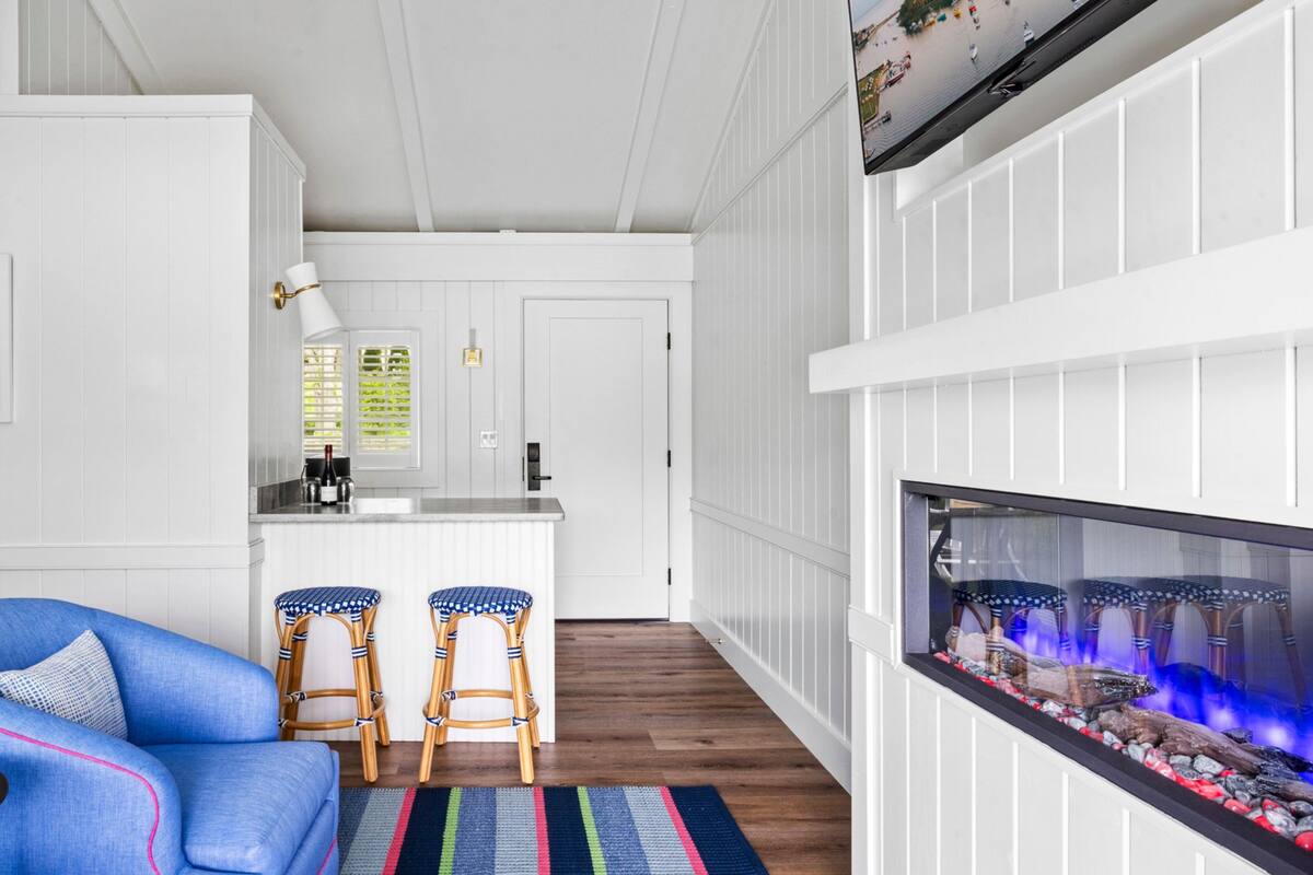 A modern living room with a blue sofa, two wicker chairs, a colorful striped rug, a fireplace, and a kitchen area with white cabinetry and a window with blinds.