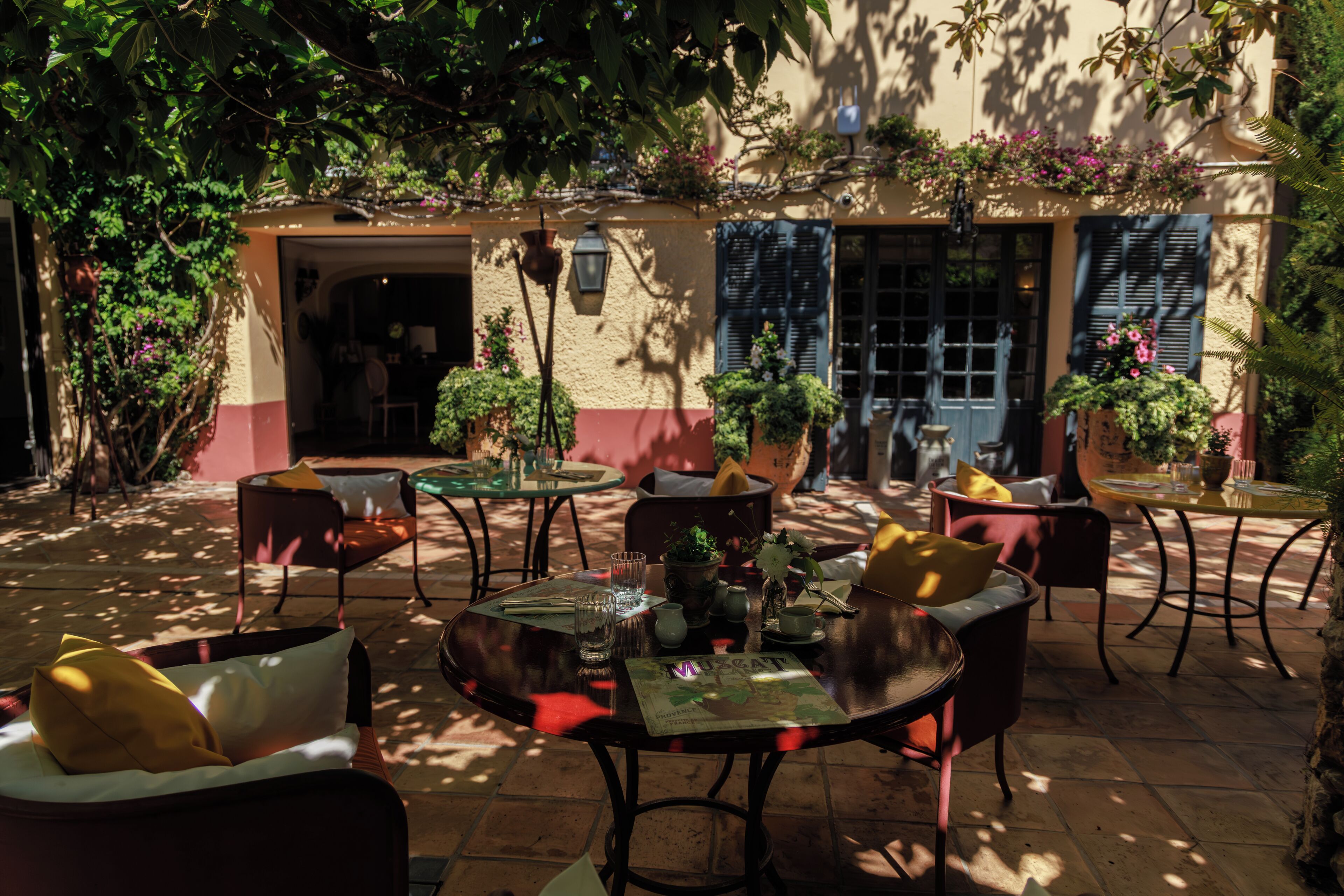 Un espace extérieur aménagé en terrasse, avec des tables et des chaises, entouré de plantes en pot et bordé d’un bâtiment doté de fenêtres et de volets.