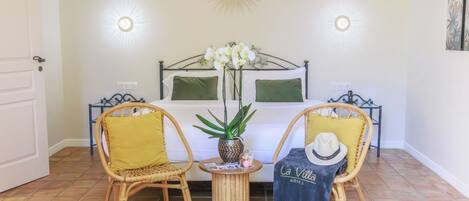 A bedroom with a bed, two chairs, a small table with a plant, and wall-mounted lights.