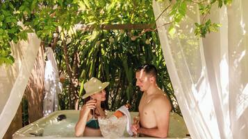 A hot tub with a wooden interior, surrounded by greenery and a white canopy.