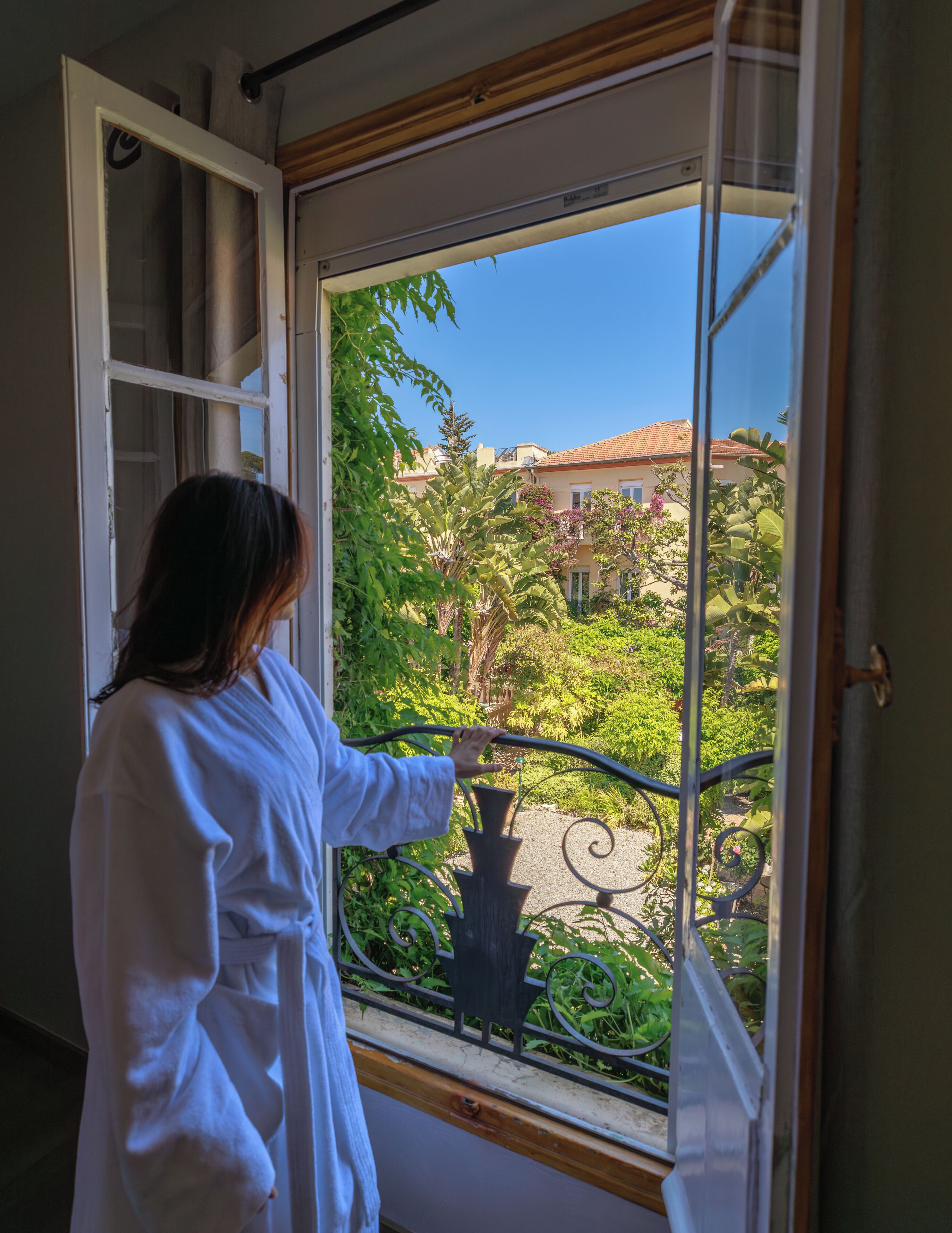 Eine Person in einem weißen Bademantel öffnet ein Fenster mit einem Metallgeländer, dahinter Blick auf ein Gebäude mit einem Ziegeldach und Grünflächen.