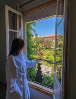 A person in a white robe opening a window with a metal railing, revealing a view of a building with a tiled roof and greenery.
