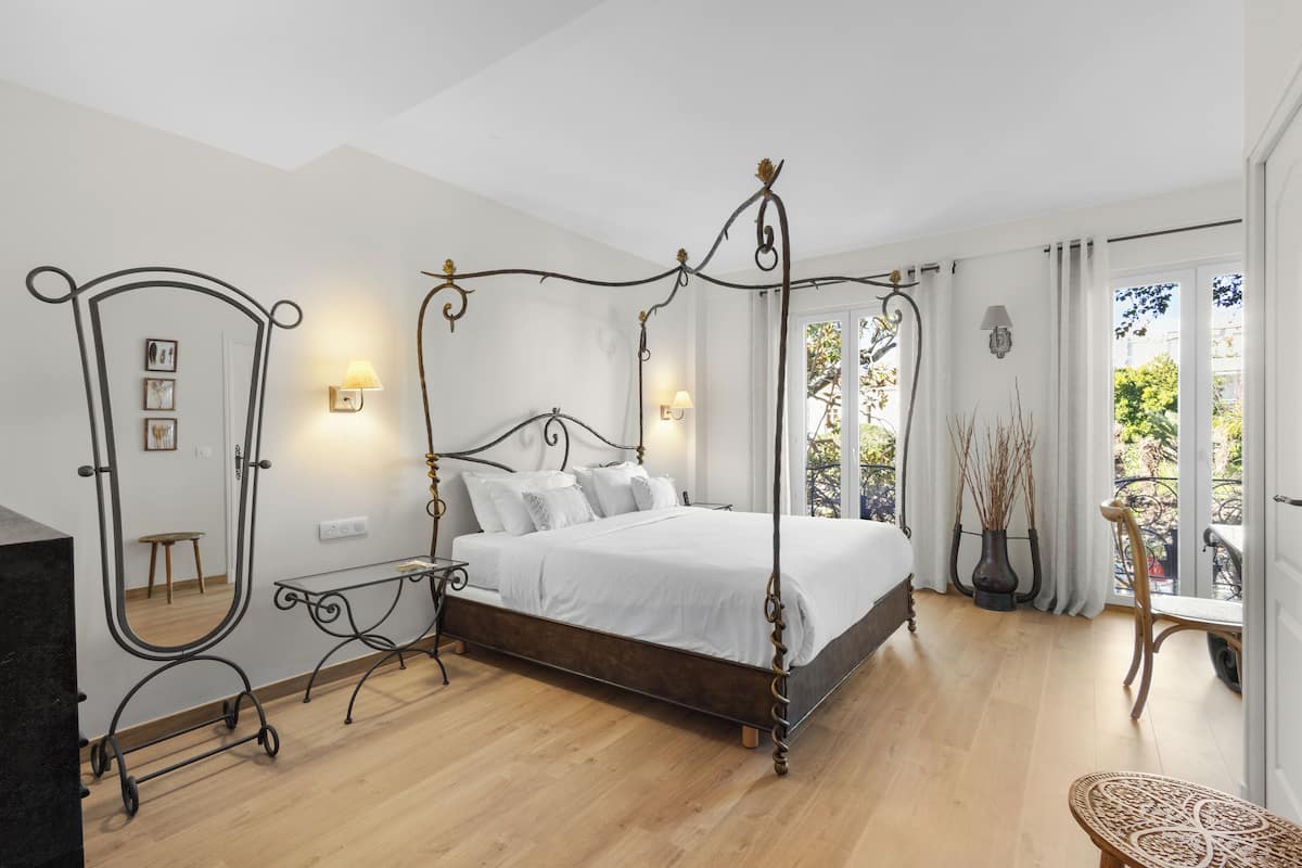 Un lit à baldaquin avec une structure en bois et une literie blanche, une petite table de chevet, un miroir et une vue sur l’extérieur par une fenêtre.