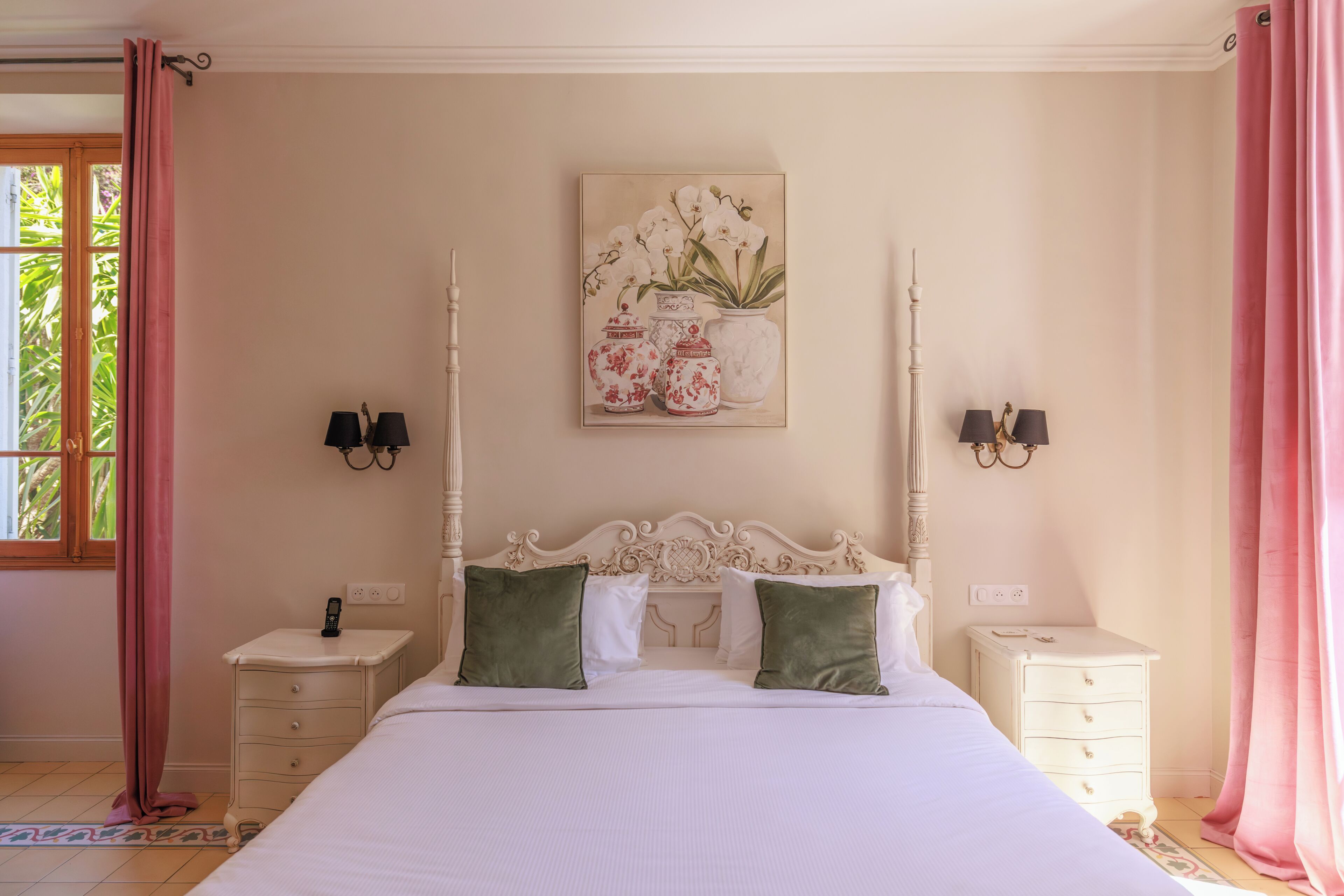 Un dormitorio con una cama grande, dos mesitas de noche, una mesita auxiliar, una lámpara de pared y una ventana con cortinas.