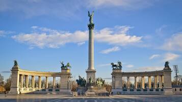 Una amplia plaza abierta con un monumento central que presenta estatuas sobre pedestales, rodeada de columnas.