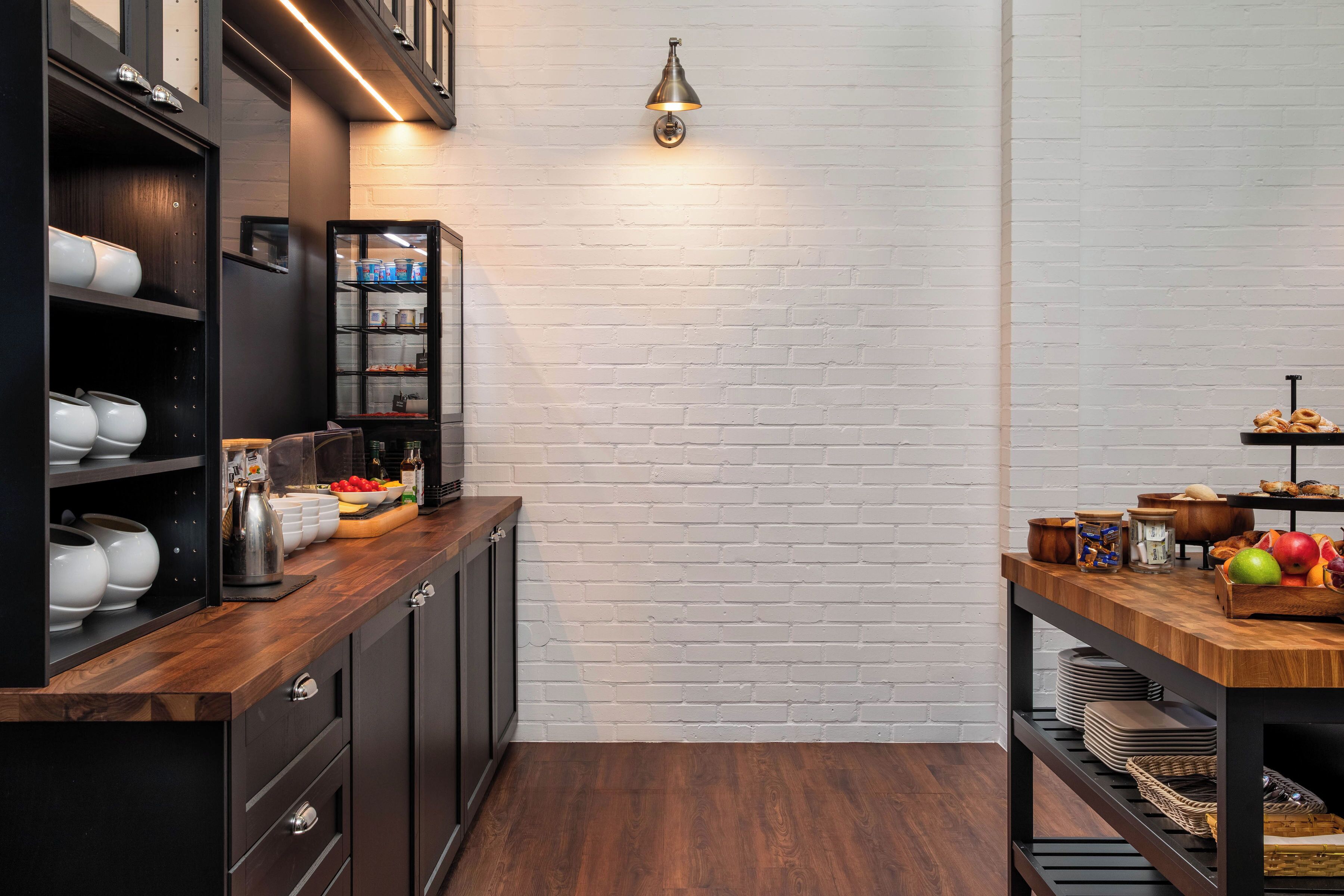 A modern kitchen with dark cabinetry, a wooden countertop, and a brick wall. There is a shelf with dishes and a small table with fruits and condiments.