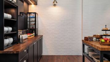 A modern kitchen with dark cabinetry, a wooden countertop, and a brick wall. There is a shelf with dishes and a small table with fruits and condiments.
