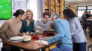 Un groupe de personnes est assis à une table dans un restaurant, et un écran diffusant des événements sportifs est visible en arrière-plan.