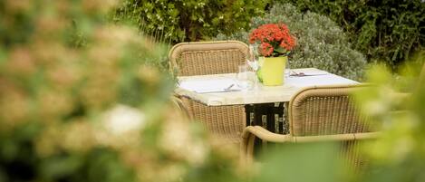 Una mesa de mimbre con un jarrón amarillo de flores rojas, una copa de vino blanco y un menú sobre un mantel blanco en un entorno al aire libre.