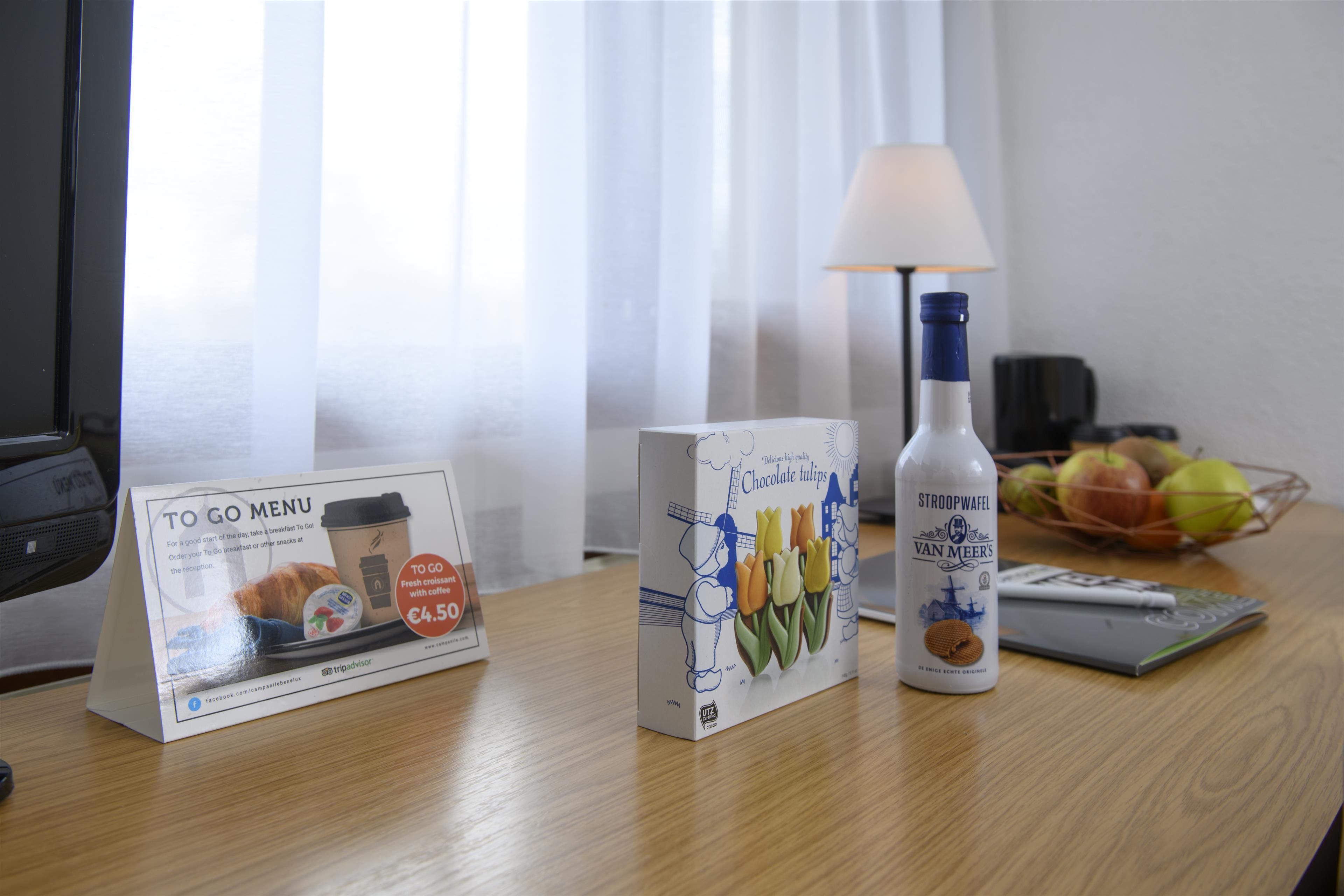 Une chambre d’hôtel avec une table en bois, un menu, une bouteille de stroopwafels et un panier de fruits.