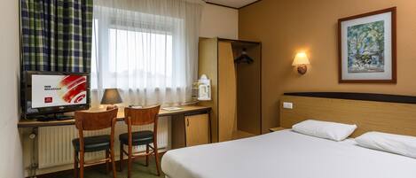 Una habitación de hotel con cama, un escritorio con computadora, una silla, un radiador, una ventana con cortinas, una lámpara de pared y un cuadro enmarcado.