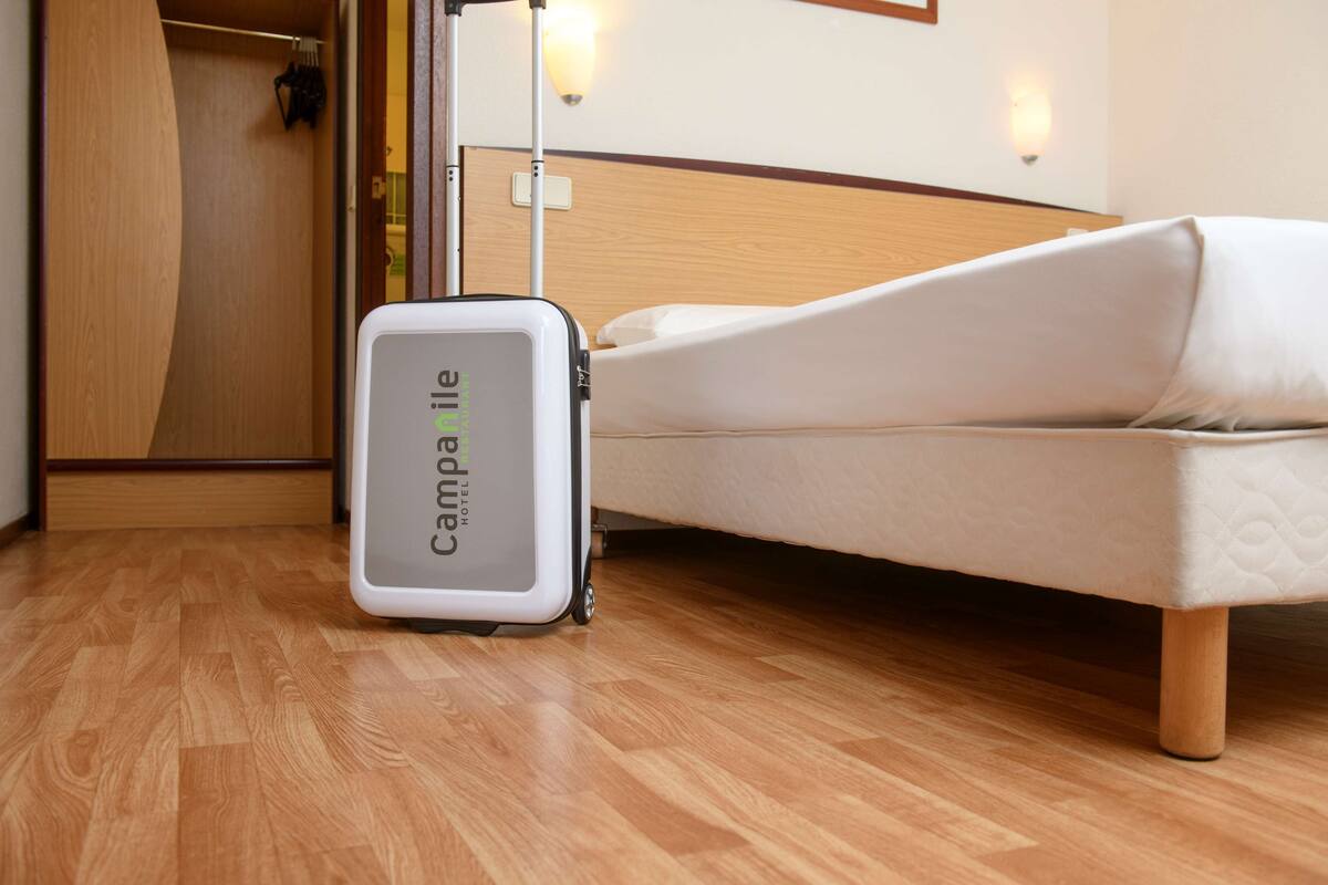 A hotel room with a bed, a wooden floor, a suitcase, and a wall-mounted light fixture.