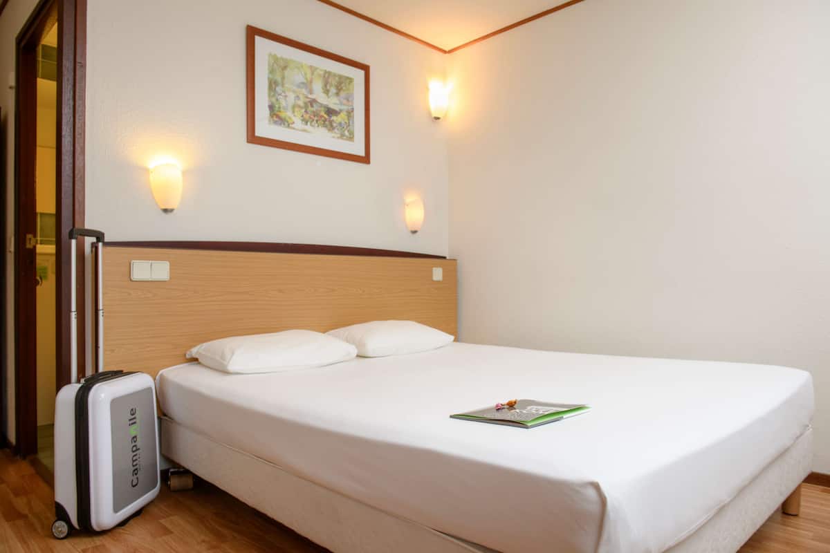 A neatly made bed with white linens, a single pillow, and a magazine on the bed. There is a suitcase on the floor to the left of the bed.
