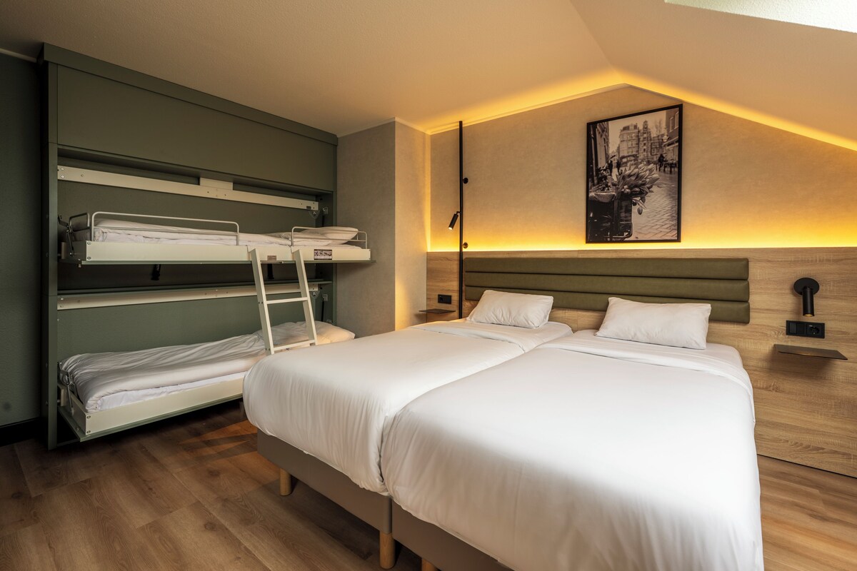 Een hotelkamer met een stapelbed, een enkel bed, houten vloeren, een wandlamp en een lijst met een afbeelding.
