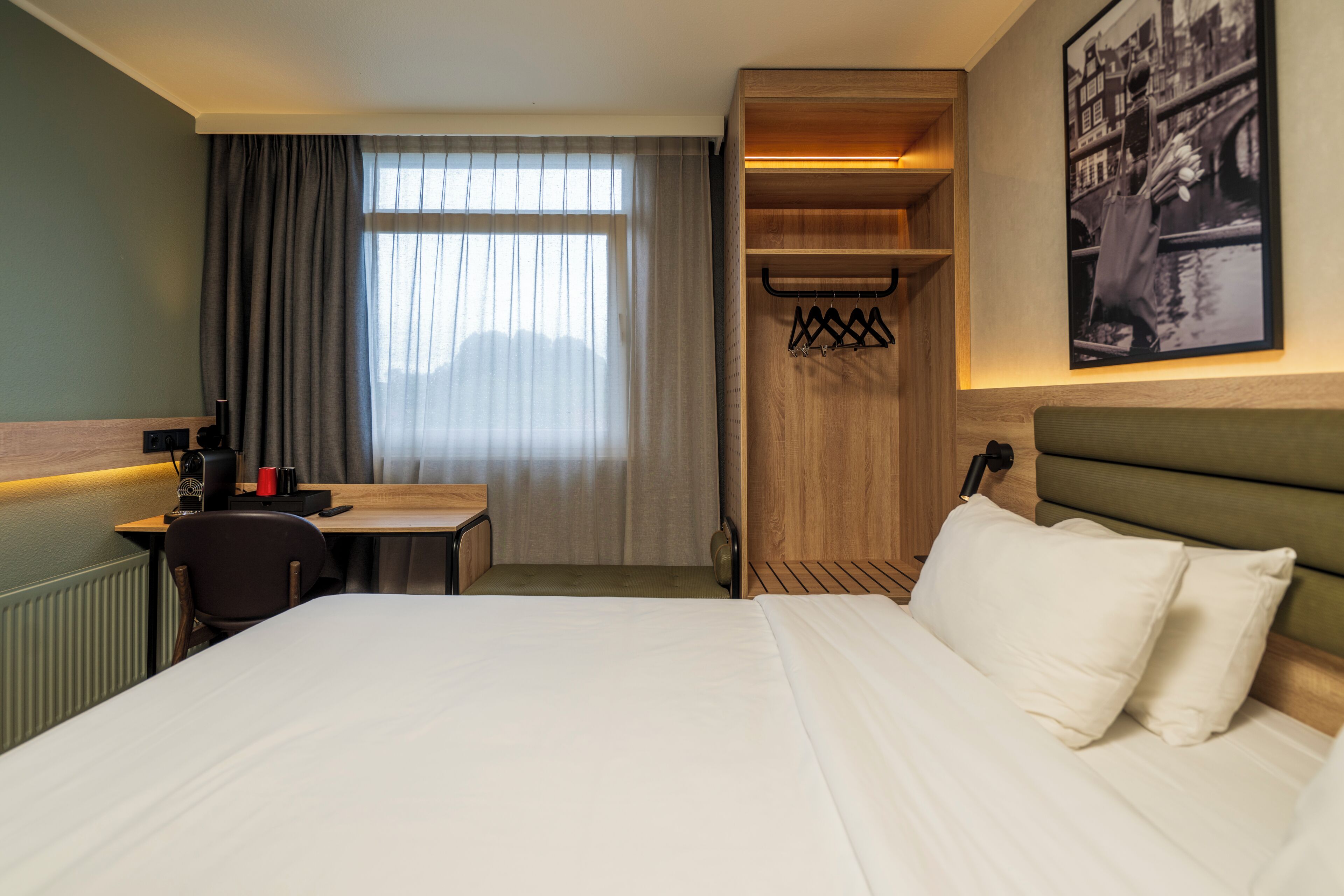 A hotel room with a bed, a desk, a chair, a coat rack, and a window with curtains.