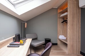 A compact room with a skylight, a desk with a remote control, a chair, a small table with a kettle, and a wooden shelving unit with folded towels. - TULIP Hotels & Residences - Zwolle (Zwolle)