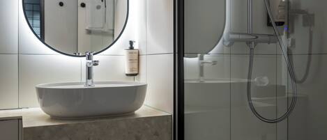 Um banheiro com uma pia oval grande, um espelho, chuveiro manual e uma garrafa de shampoo.