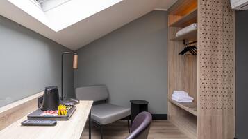 A compact room with a skylight, a desk with a coffee maker, a chair, a small round table, and a wooden wardrobe with shelves.