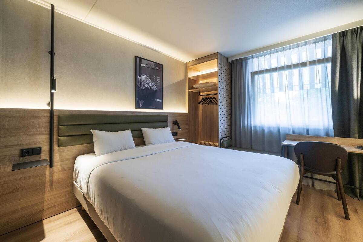 Una habitación de hotel con cama, escritorio, silla, armario y ventana con cortinas.