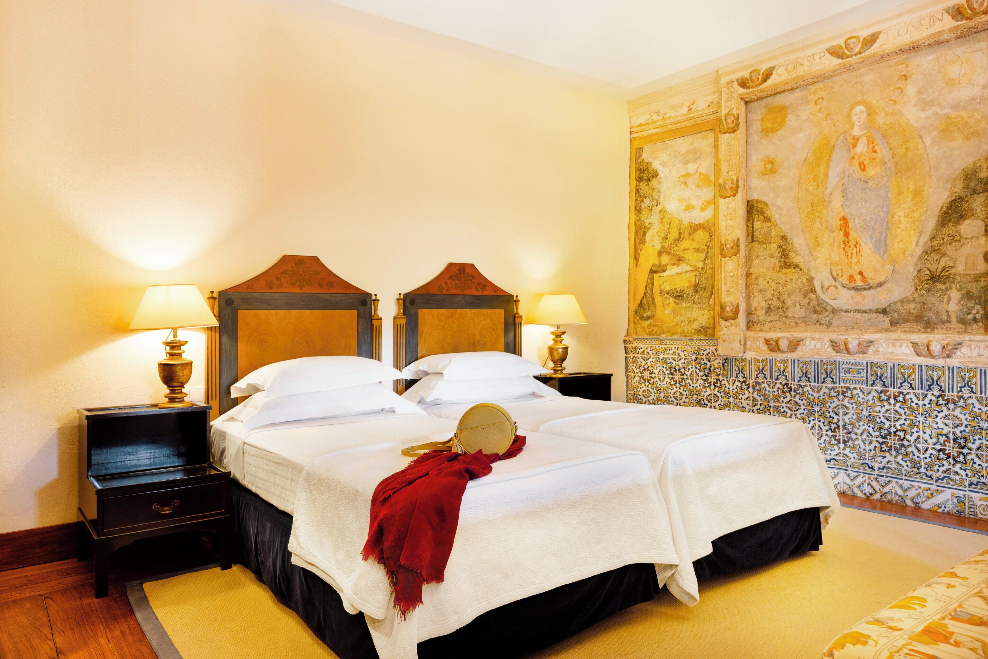 A hotel room with a large bed, two bedside tables with lamps, a red throw blanket, and a mural on the wall.