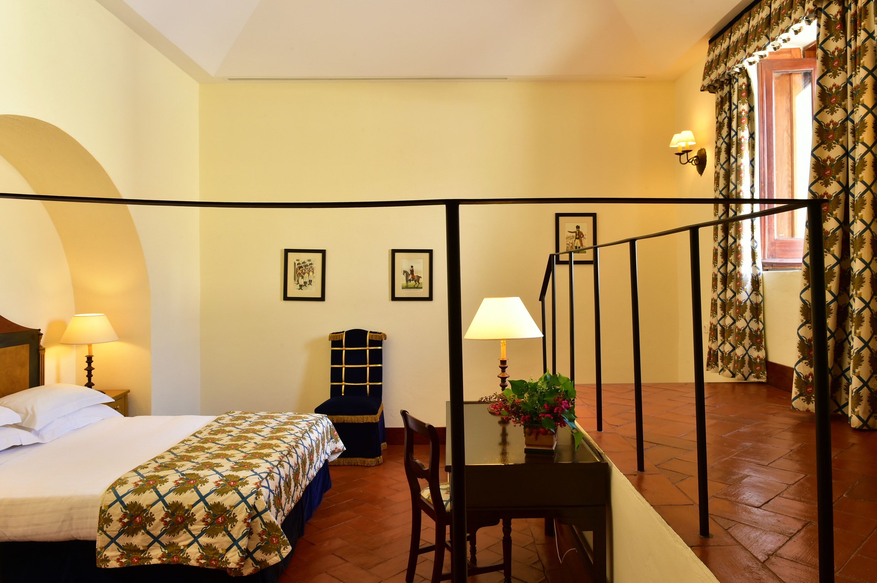 Habitación de hotel con cama, escritorio con lámpara, una silla, una mesita con planta y una escalera.