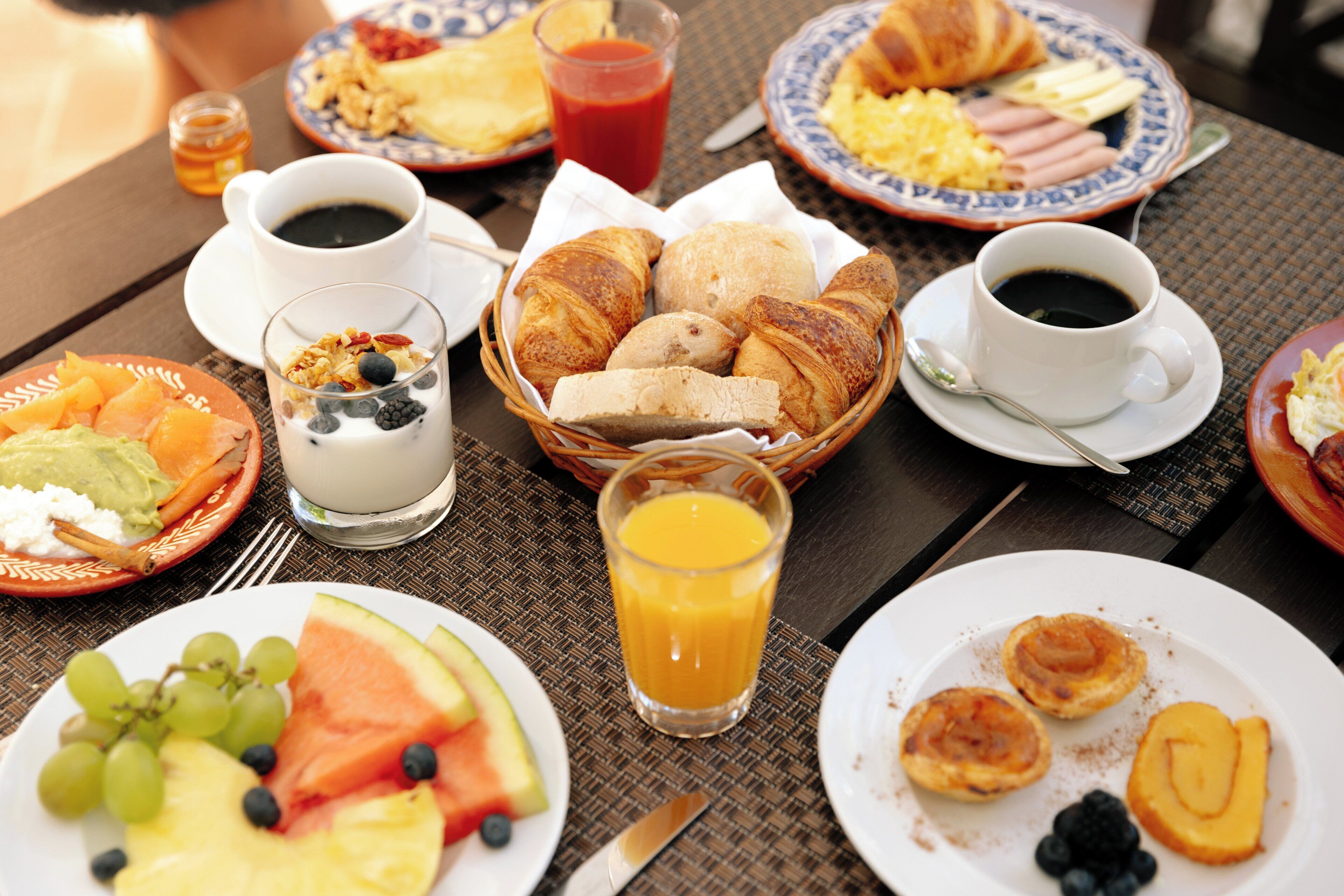 Une table de petit-déjeuner avec des assiettes remplies de nourriture, un panier de pain, un verre de jus d’orange et des tasses de café.