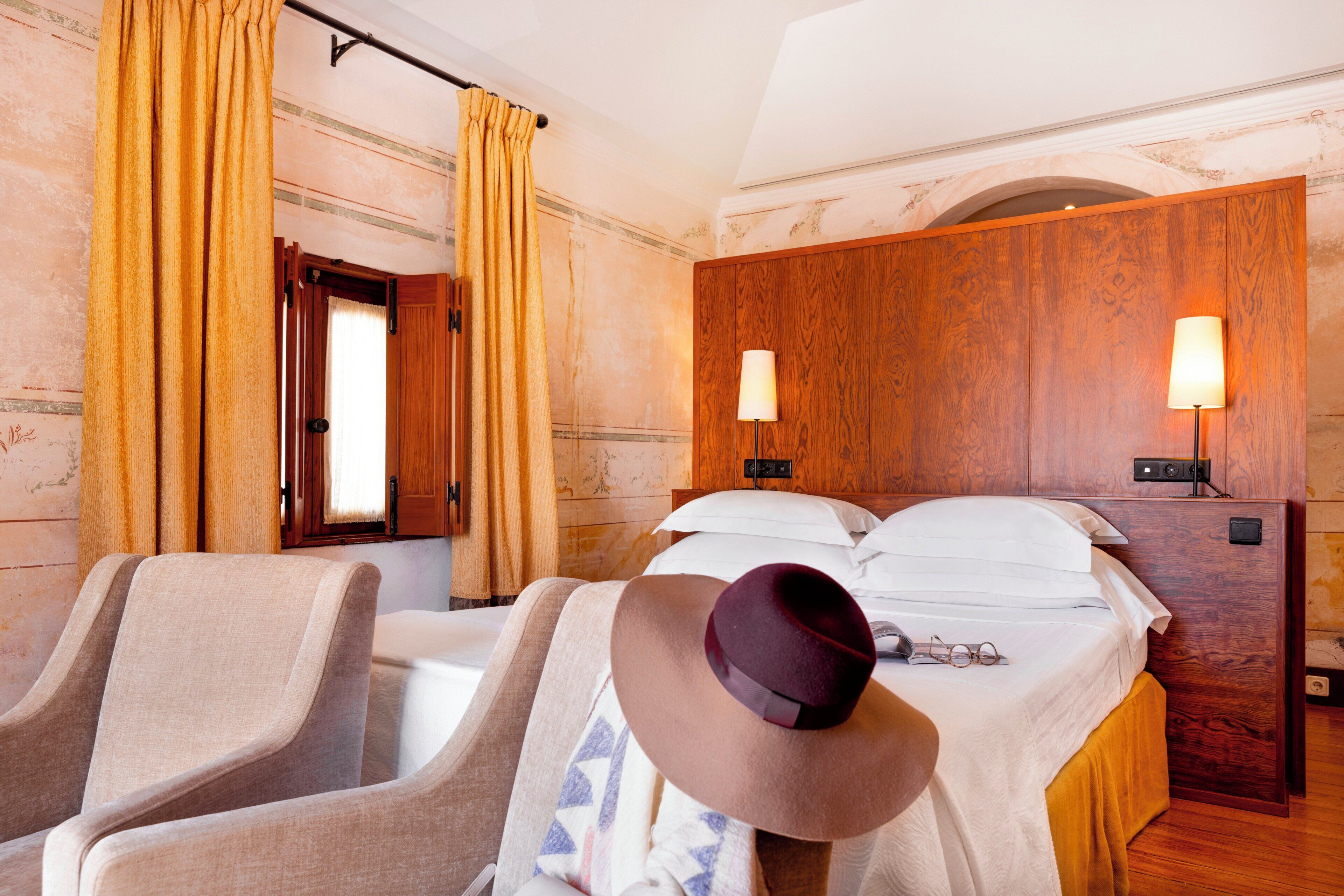 Habitación de hotel con cama, dos lámparas de noche, cabecero de madera, ventana con cortinas y un par de sillas.