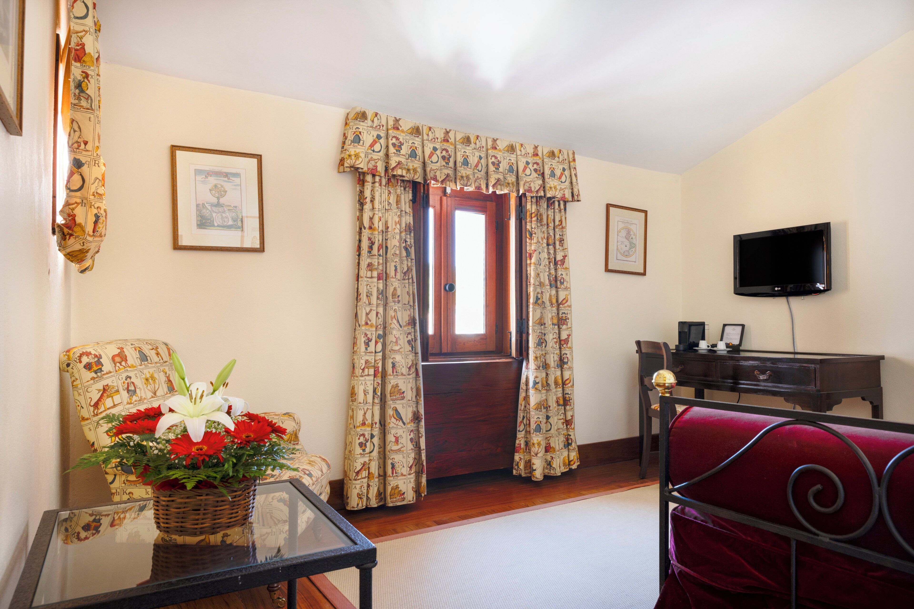 A room with a wooden door, a television, a red chair, a table with a basket of flowers, and curtains with a pattern.