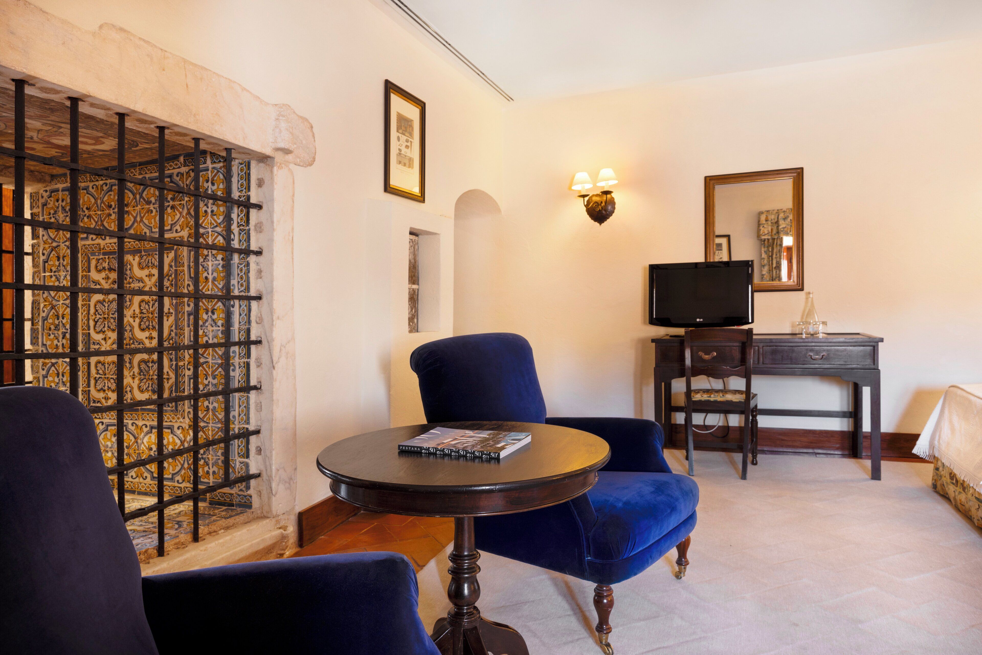 A room with a blue armchair, a small round table with a book, a television on a wooden stand, and a patterned wall.