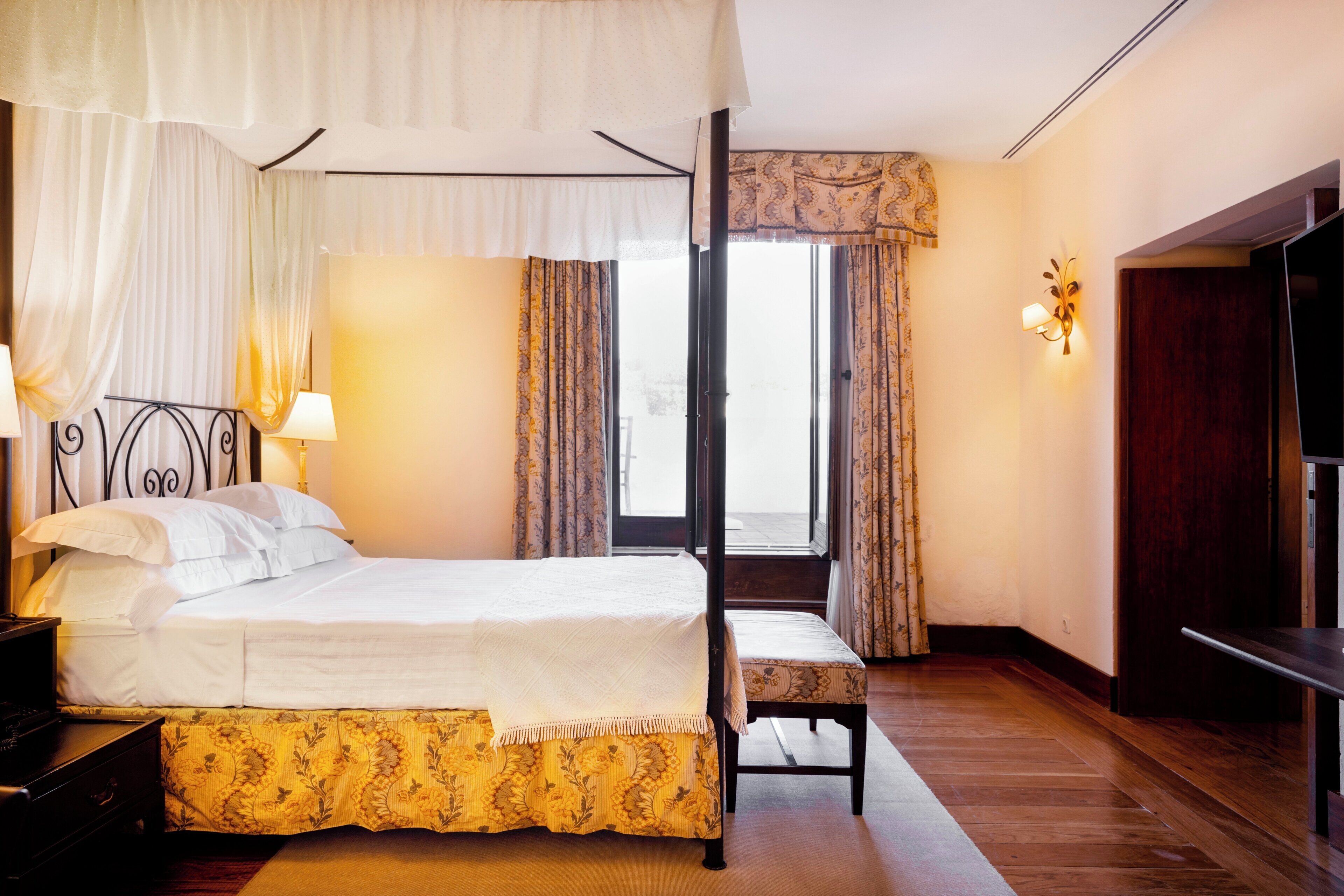 A hotel room with a four-poster bed, a bedside table, a chair, a wardrobe, and a window with curtains.